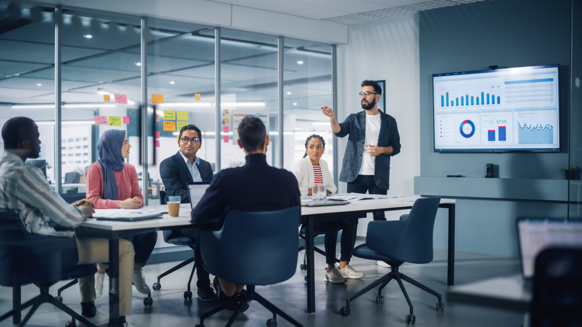 A man presents data on a large screen to five colleagues seated around a conference table in a modern office meeting room.