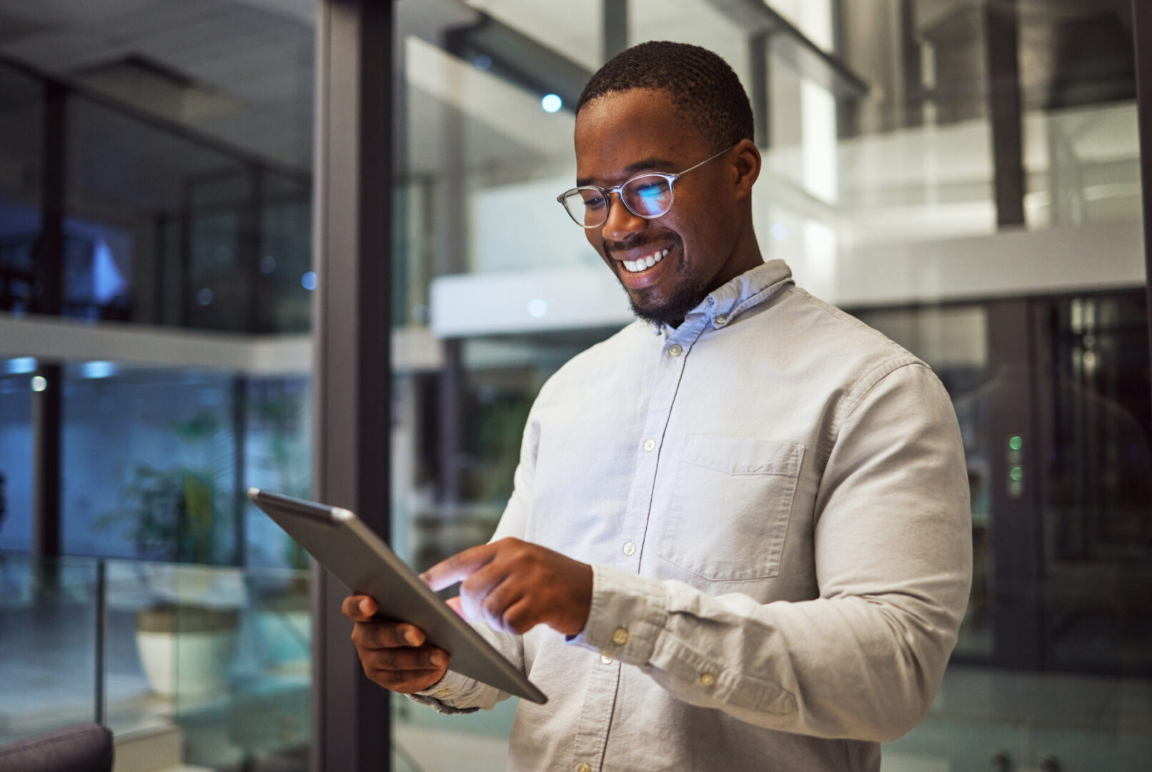 A man wearing glasses and a light shirt smiles while using a tablet in a modern office with large windows at night.