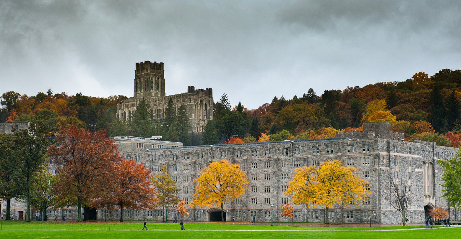 A historic castle behind an autumnal row of trees and a gray building.
